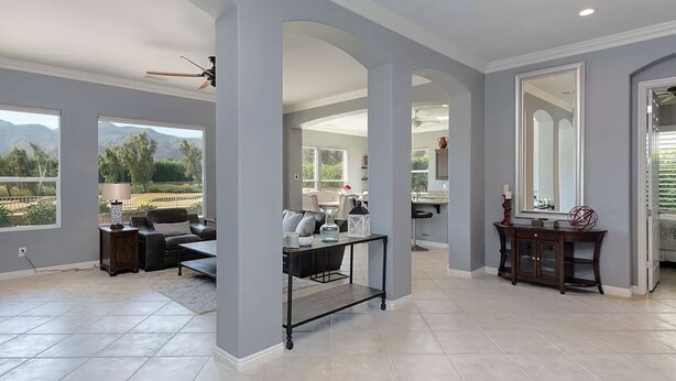 A spacious living area with a large window, a ceiling fan, and a dining area with a table and chairs. - Trilogy  Desert Oasis with Salt Water Pool and Jacuzzi with beautiful Mtn Views (La Quinta)