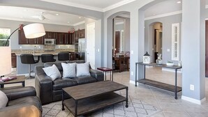 A modern living room with a dark leather sofa, a wooden coffee table, and a kitchen in the background. - Trilogy  Desert Oasis with Salt Water Pool and Jacuzzi with beautiful Mtn Views (La Quinta)