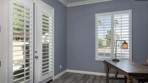 A room with a grey wall, a wooden floor, a table, and a window with blinds. - Trilogy  Desert Oasis with Salt Water Pool and Jacuzzi with beautiful Mtn Views (La Quinta)