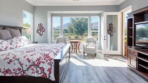 A bedroom with a bed, a TV, a dining table with chairs, and a view of the outdoors. - Trilogy  Desert Oasis with Salt Water Pool and Jacuzzi with beautiful Mtn Views (La Quinta)