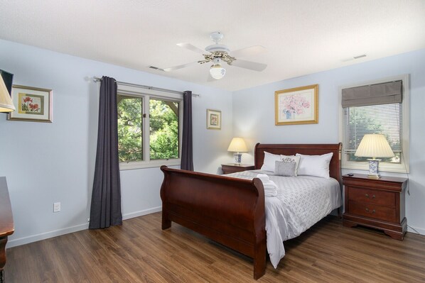 A bedroom with a wooden bed, two bedside tables with lamps, a ceiling fan, and a window with curtains.