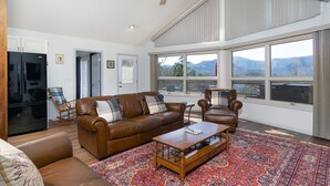 Ein geräumiges Wohnzimmer mit einem braunen Ledersofa, einem Holztisch und einem gemusterten Teppich. Ein großes Fenster bietet Blick auf die Berge.