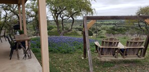 A rustic outdoor seating area with a wooden table and chairs, surrounded by a lush garden and a scenic view of rolling hills. - The Cowhouse, a cozy hilltop guesthouse at Barkspin Oaks Ranch. Beautiful views! (Dripping Springs)