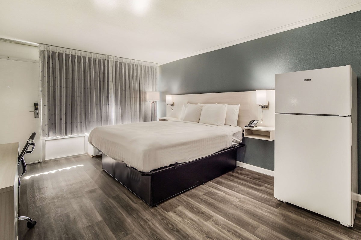 Een hotelkamer met een bed, een koelkast, een nachtkastje, een lamp en een raam met gordijnen.