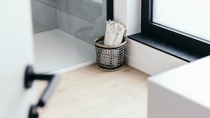 A corner of a room with a woven basket containing towels, a wooden floor, and a window with a black frame. - Factory Hotel (Beveren)