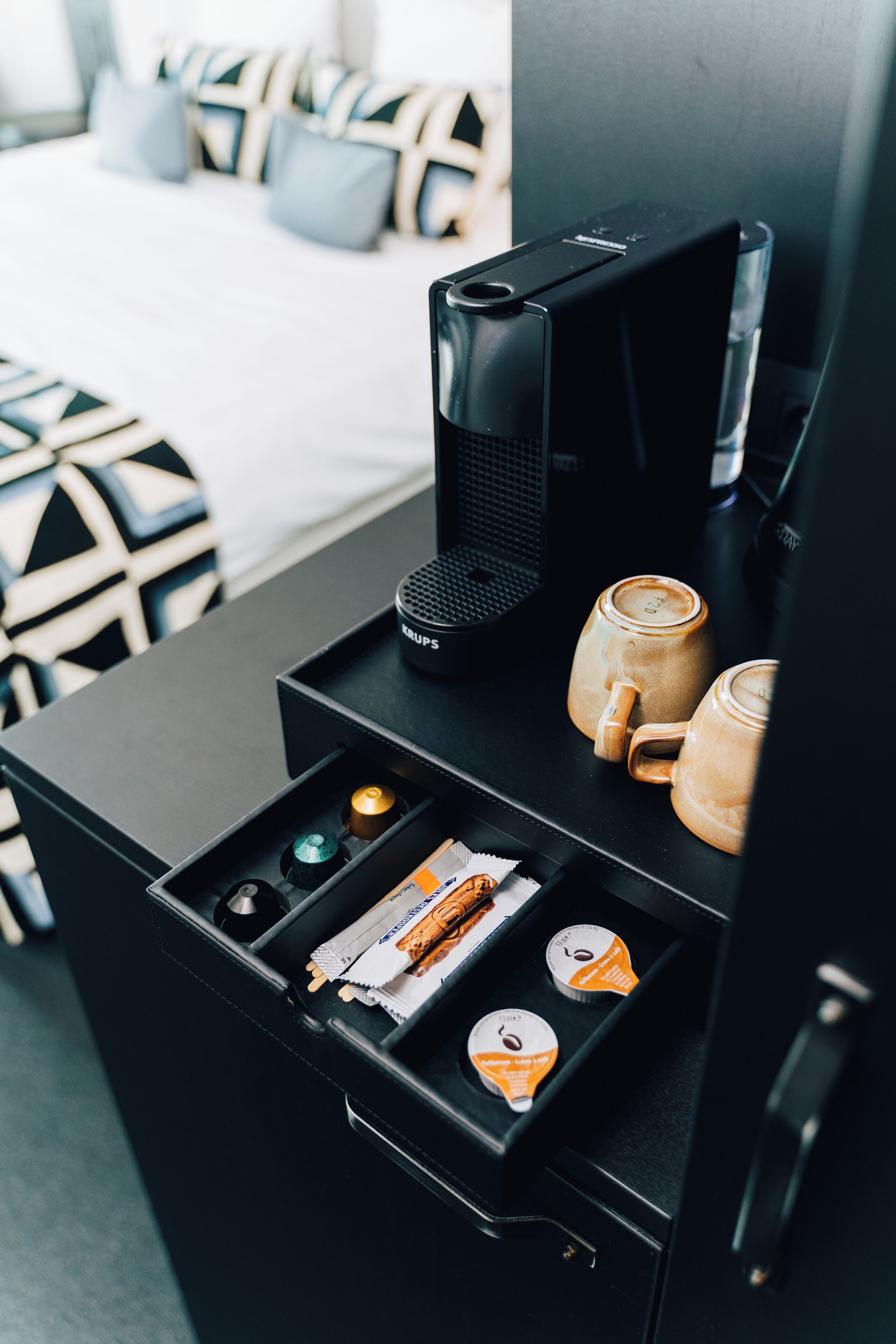 Une chambre d’hôtel équipée d’un mini-bar noir, d’une machine à café, de deux tasses à café et d’un plateau avec des en-cas et des dosettes de café.