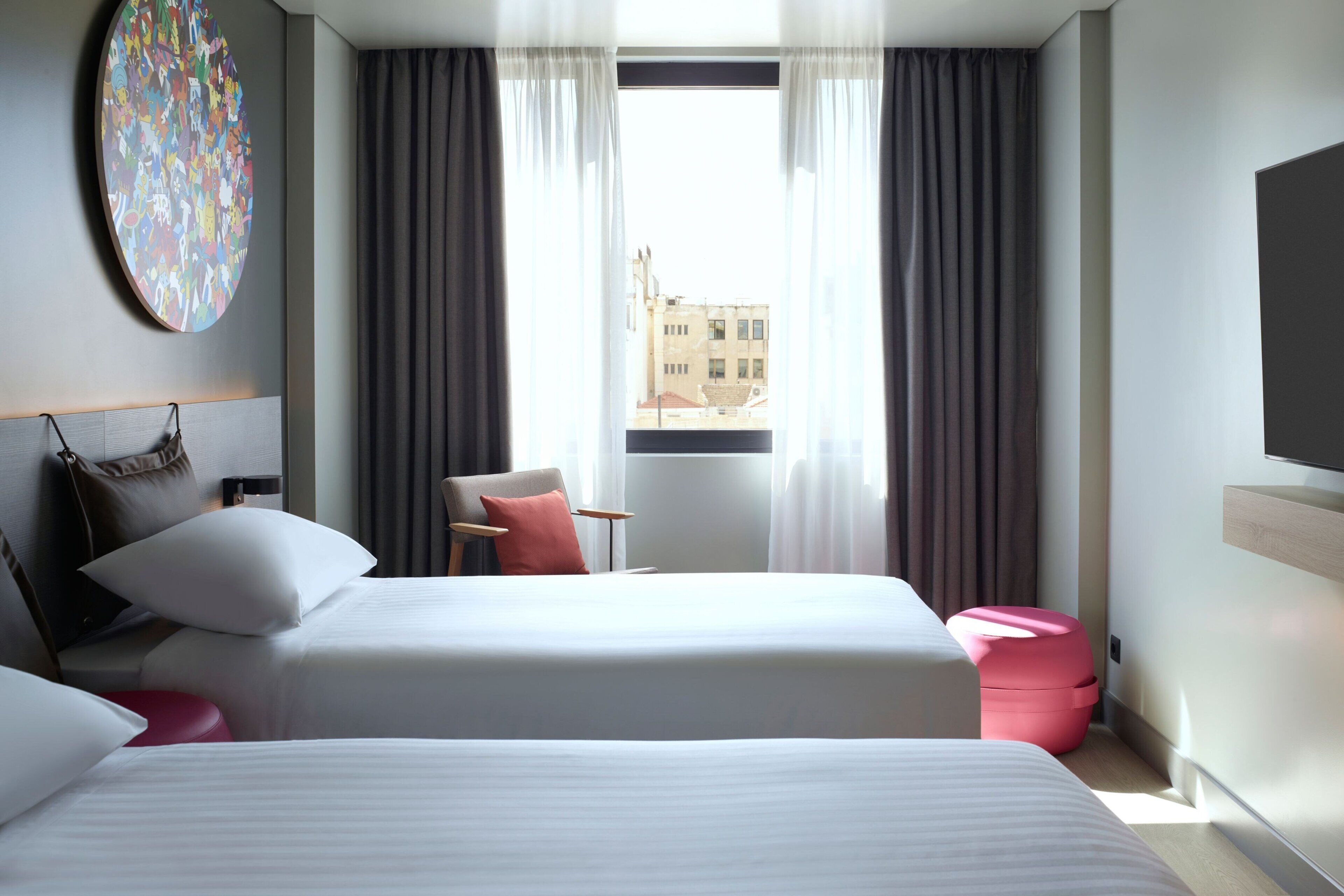 A modern hotel room with a large bed, a desk, and a chair. There is a round artwork on the wall and a view of buildings outside the window.
