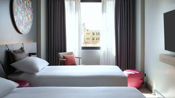 A modern hotel room with a large bed, a desk, and a chair. There is a round artwork on the wall and a view of buildings outside the window.