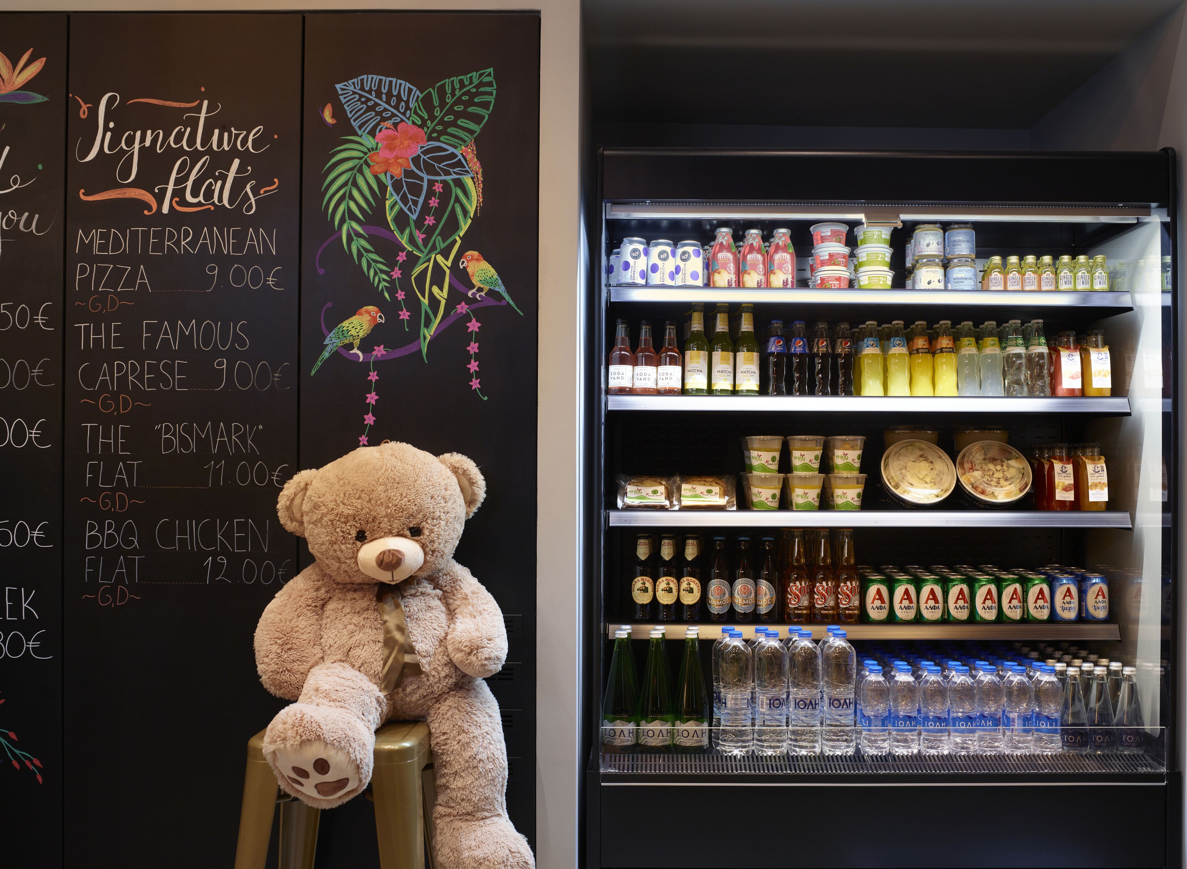 A menu board with various food items and prices, a teddy bear on a stool, and a beverage refrigerator stocked with drinks.