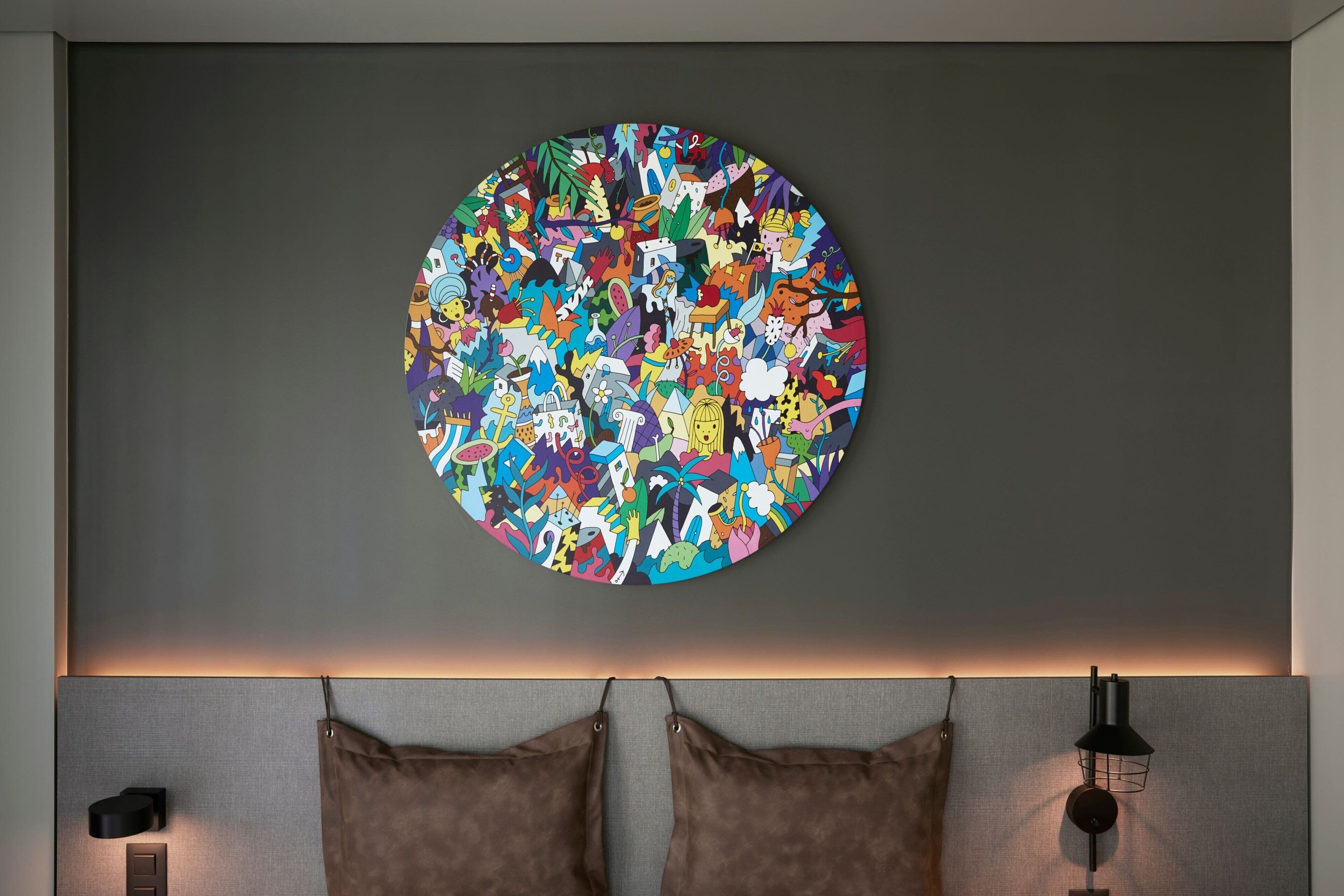 Una obra d'art circular amb diverses figures abstractes i patrons sobre una paret gris, sobre un llit amb dos almoiners marrons i un capçal.