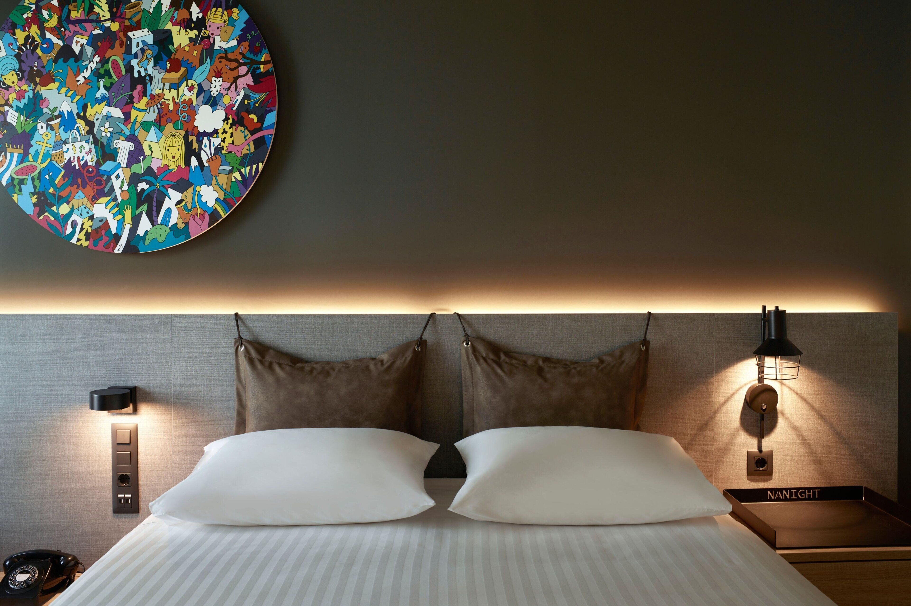 Una habitación de hotel con una cama, dos mesitas de noche, una lámpara de pared, un teléfono y una obra de arte circular sobre la cama.