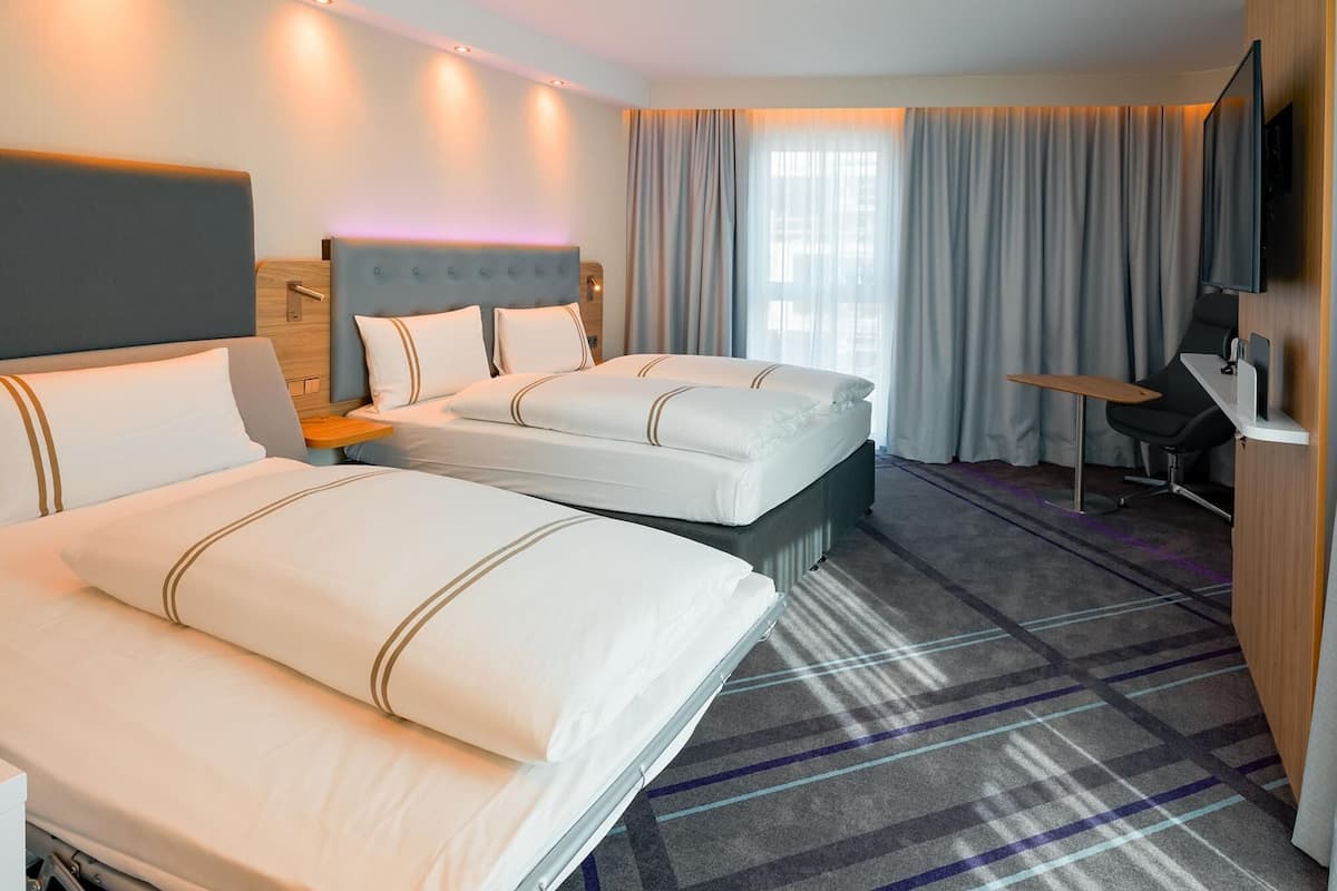 Ein Hotelzimmer mit zwei Betten, einem Schreibtisch, einem Sessel, einem Fernseher und einem großen Fenster mit Vorhängen.