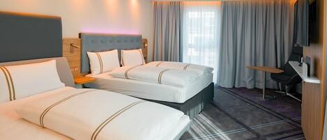 A hotel room with two beds, a desk, a chair, a TV, and a large window with curtains.