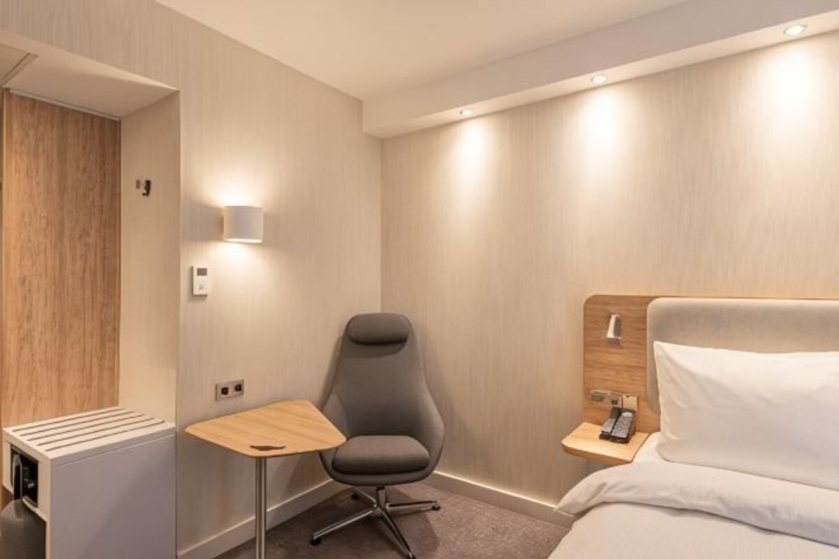 A hotel room with a bed, a chair, a small table, a bedside table with a lamp, and a wall-mounted heater.