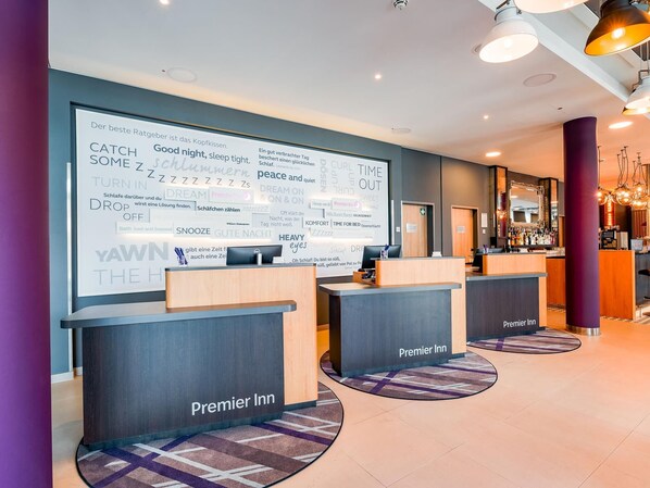 A hotel reception area with a large wall displaying various sleep-related words and phrases, reception desks labeled 'Premier Inn,' and a bar area in the background.