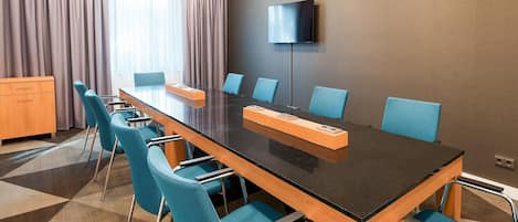A conference room with a long table, blue chairs, a flat-screen TV, and a window with curtains.