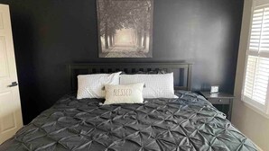 A bedroom with a dark quilted bedspread, a headboard, and a wall-mounted picture of a forest path.