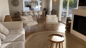 A spacious living room with a fireplace, a dining area with a round table and chairs, and a large window providing a view of the outdoors. - Magnificent villa with swimming pool in the center of Cap Ferret (CAP FERRET)