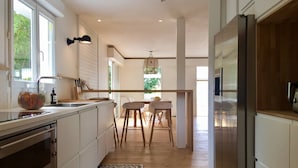 A modern kitchen with a central island, a dining area with a wooden table and chairs, and a view of greenery outside. - Magnificent villa with swimming pool in the center of Cap Ferret (CAP FERRET)
