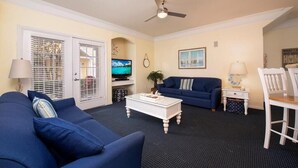 Ein Wohnzimmer mit blauem Sofa, weißem Couchtisch, Fernseher und Deckenventilator.