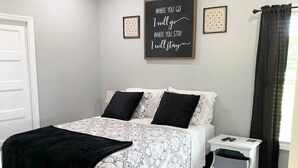 Een slaapkamer met een bed, een nachtkastje, een plafondventilator, een zwart-wit beddekleed, een zwarte plaid, een vloerkleed en een motiverende quote op de muur.