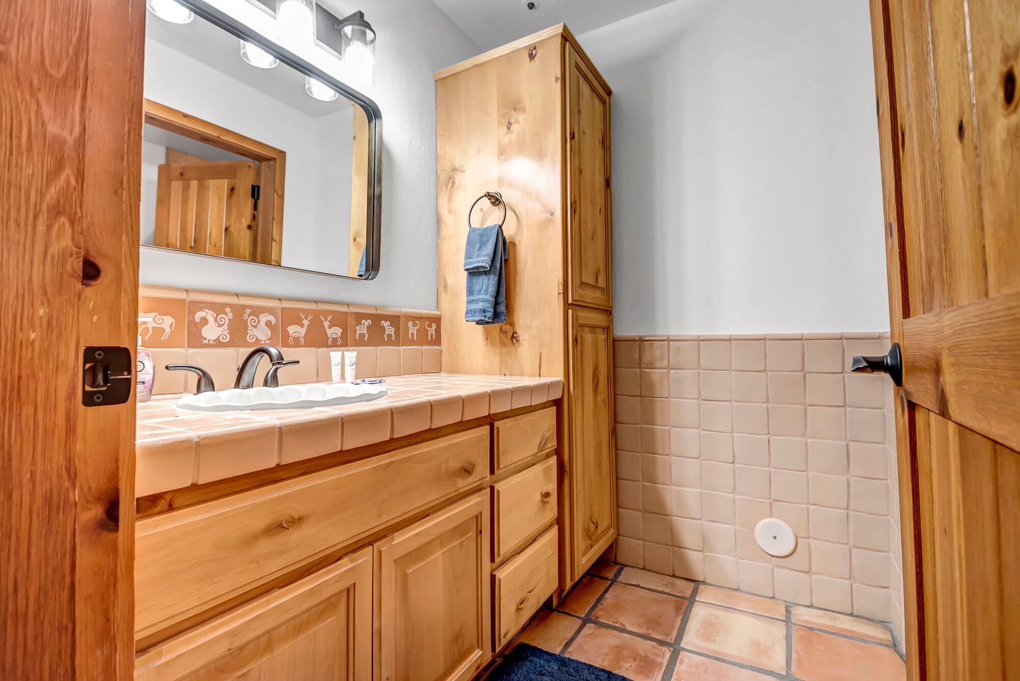 A bathroom with a wooden vanity, a sink, and a mirror. There are decorative tiles on the wall and a towel hanging on the door.