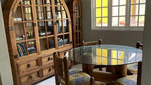 Un comedor con una mesa redonda de vidrio, sillas de madera y un aparador de madera con puertas de cristal, además de una ventana con persianas amarillas.