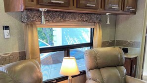 Un interior de autocaravana acogedor con dos sillones de cuero reclinables, una lámpara de mesa y armarios de madera sobre un ventanal.