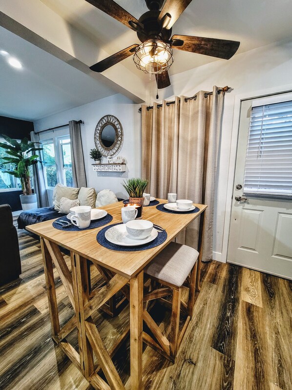 A dining area with a wooden table set for four, a ceiling fan, and a view of the outdoors through a window.
