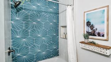 A modern bathroom with a geometric tile shower wall, a white bathtub, a wooden floor, and a framed picture on the wall.