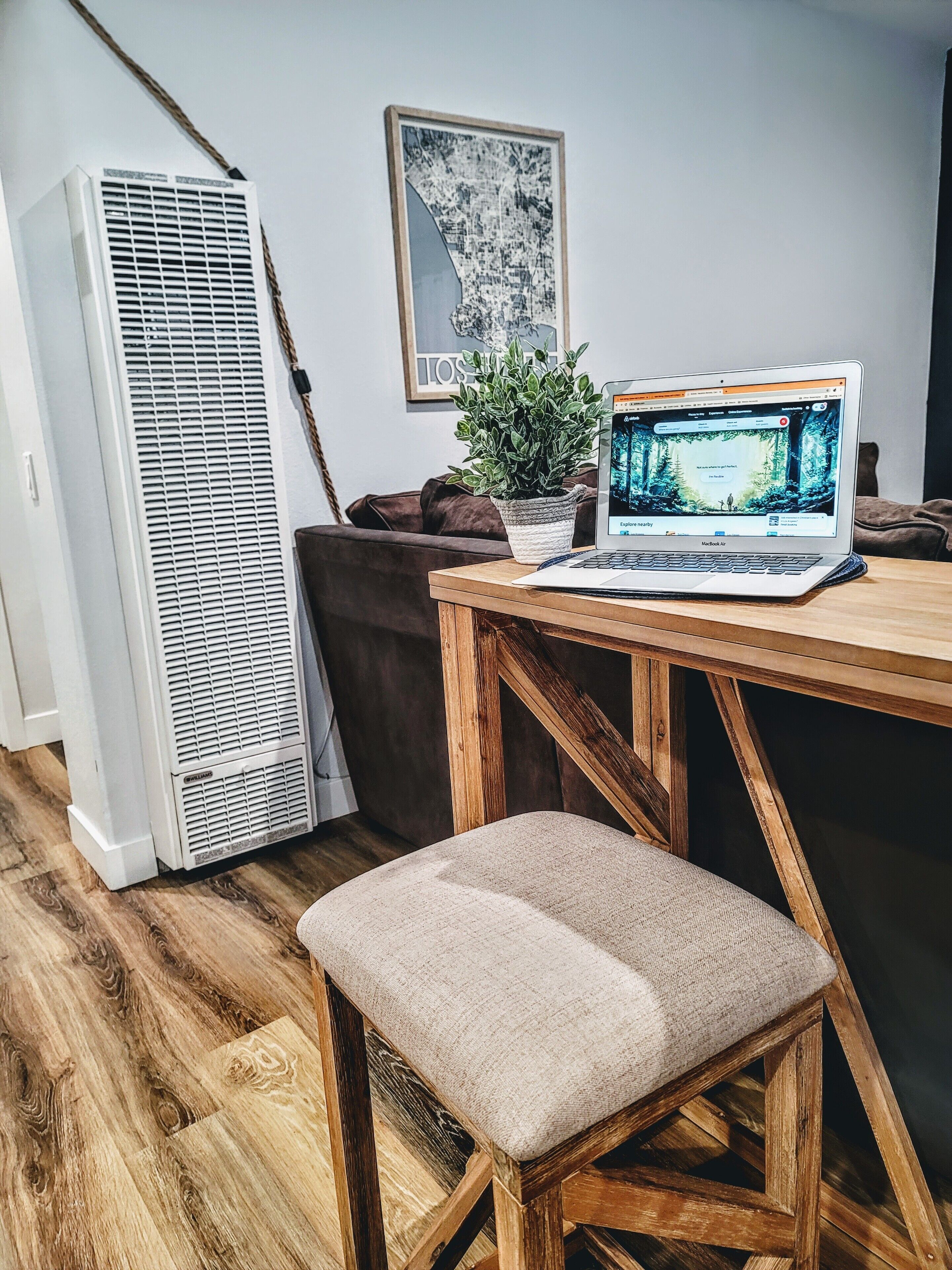 A room with a wooden table, a laptop, a potted plant, a tall air conditioner, and a framed picture on the wall.