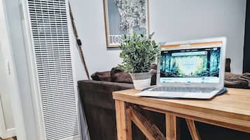 A room with a wooden table, a laptop, a potted plant, a tall air conditioner, and a framed picture on the wall.