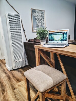 Una habitación con una mesa de madera, una computadora portátil, una planta en maceta, un aire acondicionado alto y un cuadro en la pared.
