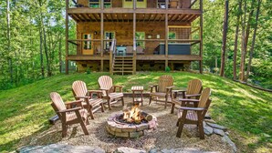 Una cabaña de madera de varios niveles con un hoyo para el fuego y sillas Adirondack en el patio.