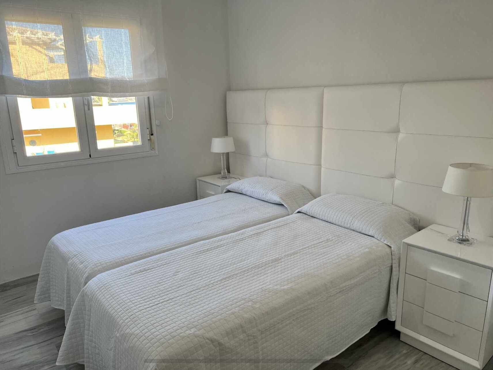 Una habitación con dos camas, una ventana y una mesita de noche con lámpara.