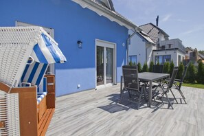 A patio with a table and chairs, a wicker chair with a striped umbrella, and a blue building with a sliding glass door. - Vacation home Luna - Holiday home Luna semi-detached house (Breege)