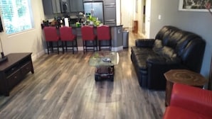 Un salon avec un canapé en cuir noir, une table basse en verre et un fauteuil rouge. On y trouve également un coin cuisine avec des chaises rouges et un réfrigérateur à l’arrière-plan.
