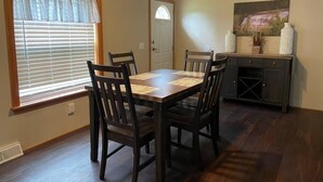 Comedor con mesa de madera y cuatro sillas, una ventana con persianas, una puerta y un mueble con objetos decorativos.