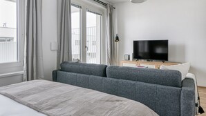 Une chambre moderne avec un grand lit, un canapé gris, une télévision et un balcon agrémenté de rideaux.