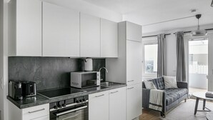 Une cuisine moderne avec des armoires blanches, un comptoir noir, un four encastré, un four à micro-ondes et une machine à café. Le salon adjacent est doté d  ’un canapé gris et d’une petite table ronde, le tout agrémenté d’un tapis.
