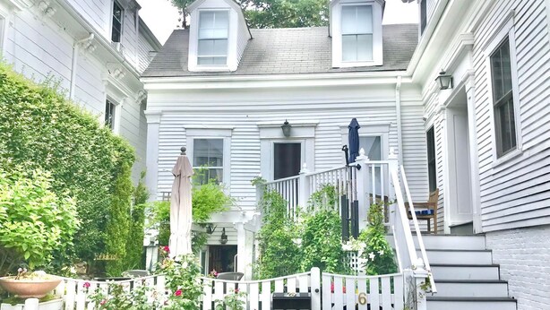 A two-story house with a white exterior, a front porch with a bench, and a small garden area with potted plants and flowers.