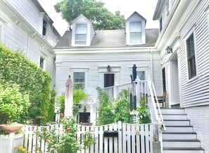 Una casa de dos pisos con fachada blanca, un porche con banco y un pequeño jardín con plantas y flores en macetas.