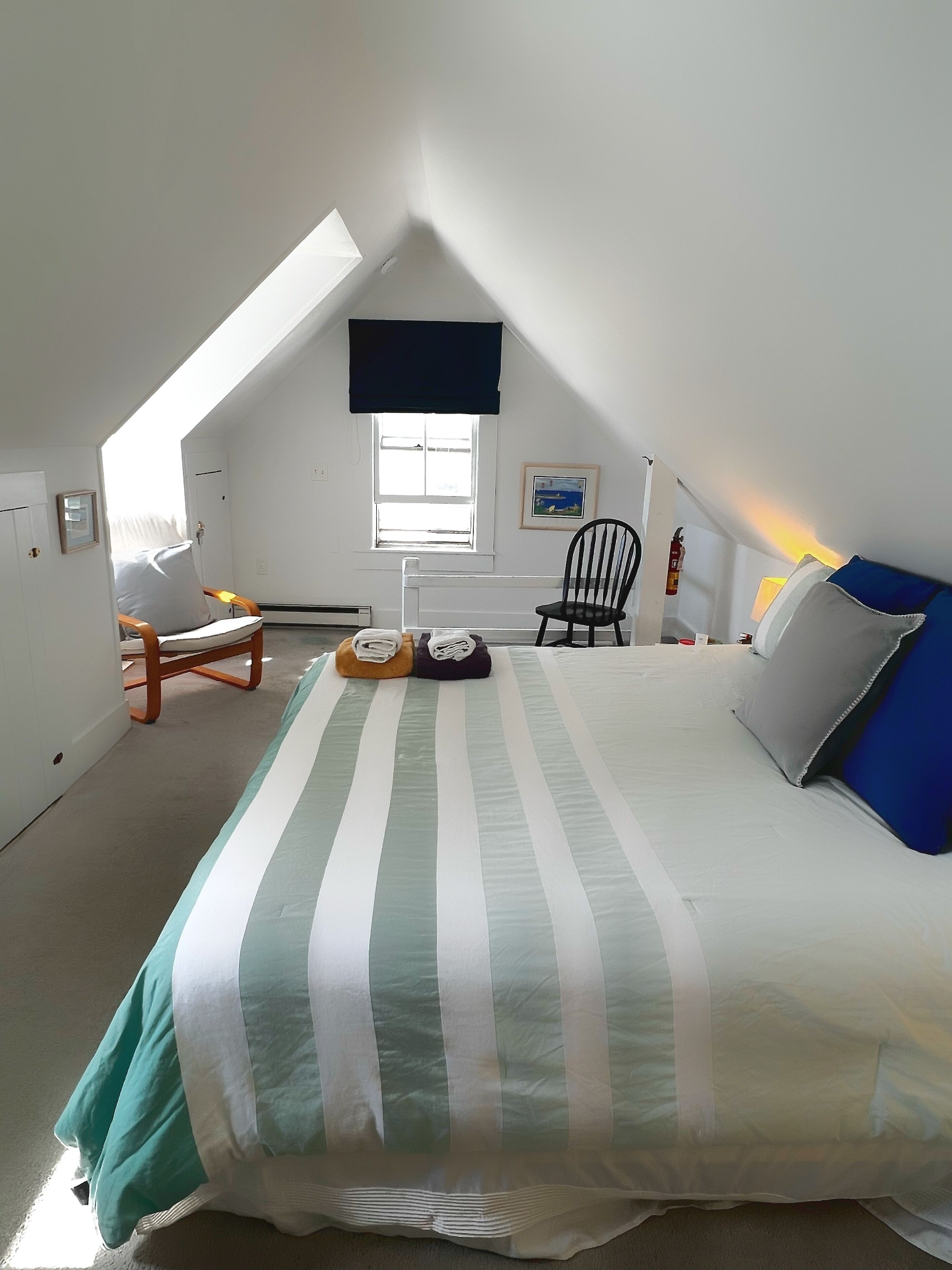 A bedroom with a sloped ceiling, a bed with striped bedding, a chair, a small table, and a window with a view.