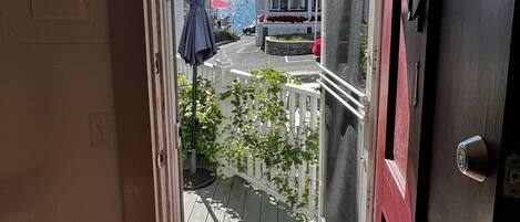 Vista a través de una puerta que da a un balcón con piso de madera, cerca hay una valla de piquetes blancos y una puerta roja.