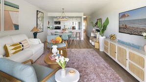 A modern living room with a sofa, chairs, a coffee table, and a dining area. There is a kitchen in the background with white cabinets and a dining table.