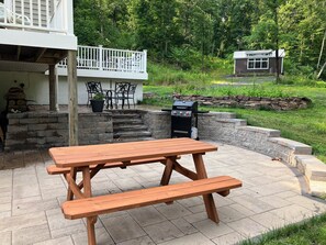 A wooden picnic table with benches in a backyard patio area with stone steps and a barbecue grill. - Creekside Retreat-spacious cottage nestled in the wildflowers and lush woods (Harleysville)