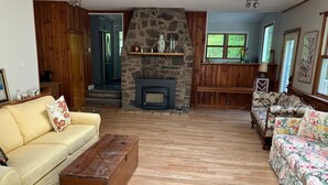 Ein gemütliches Wohnzimmer mit Kamin aus Stein, Holzmöbeln und Blick ins Freie durch große Fenster.
