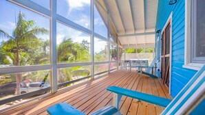 Un porche cubierto con piso de madera y barandillas azules, que da a un jardín tropical con palmeras y un automóvil.