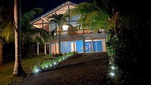 Una casa de dos pisos con un balcón, rodeada de palmeras y un camino de entrada bien iluminado.
