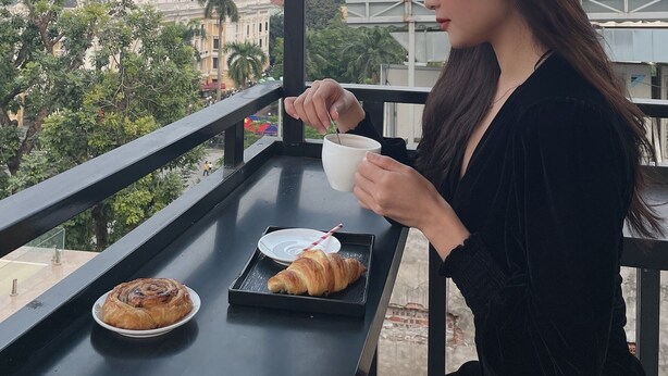 A person sitting at a table with a croissant and a cup, overlooking a cityscape with buildings and trees. - Opera House Suites (Hanoi)